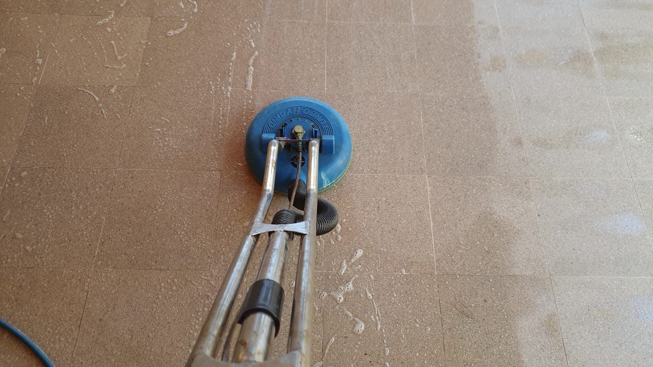 A round tile-and-grout cleaning head on beige floor tiles, the scrubbed patch paler than the untouched grout lines